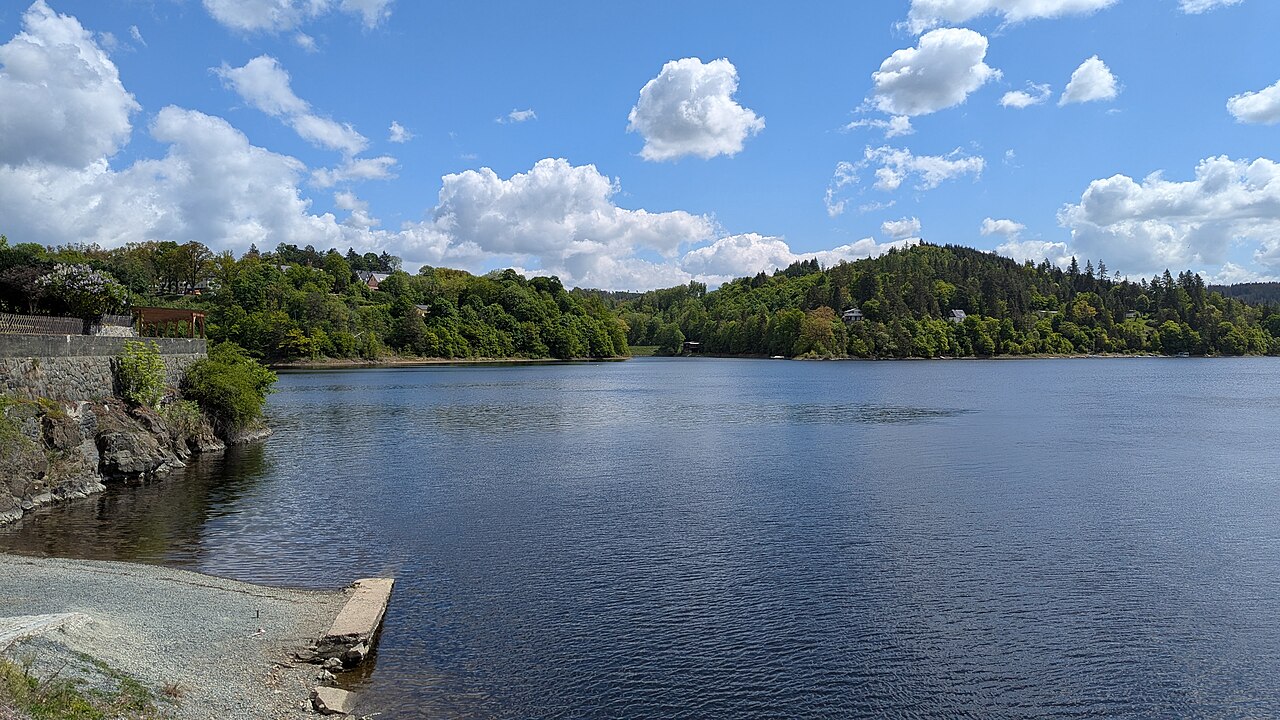 Blick vom Ufer auf die von Wald umgebende Talsperre. Links das Ufer mit dem Uferweg – Campingplatz Saalburg - Bad